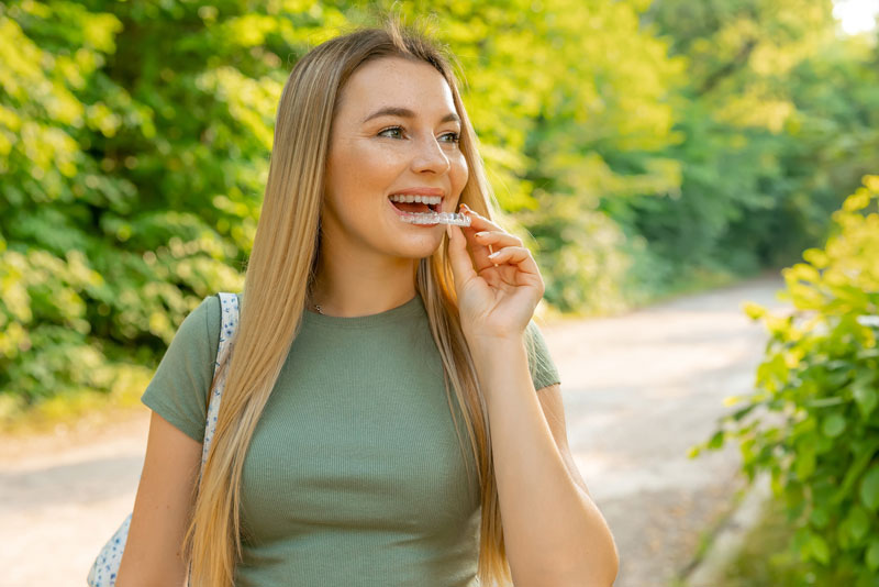 Smiling young woman outdoors holding a clear aligner near her mouth, preparing to place it on her teeth.