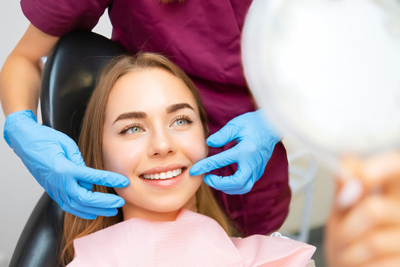 Happy dental patient in chair smiling and checking her teeth in a mirror while a dentist in gloves examines her results after cosmetic treatment.